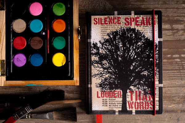 A hardcover diary with a black tree silhouette lies on a rustic wooden surface next to an open watercolor palette and paintbrushes.