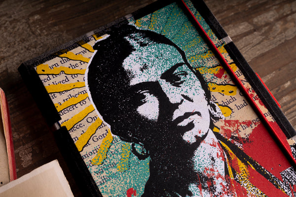 A close-up of a square decorative art piece featuring a stylized portrait  in black, white, and teal, layered over yellow book-textured panels with red accents, photographed on a wooden surface.