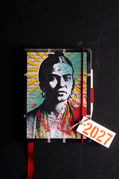 A hardcover journal featuring a stylized pop-art portrait  in a mosaic print, with a red elastic closure and a white paper tag tied to the spine that reads '2027'. The journal is photographed flat on a dark, textured background.