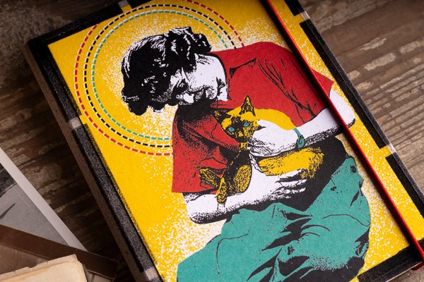 A square framed diary cover featuring a stylized illustration of a woman in a red top and teal skirt holding a yellow cat, with a bright yellow background and dotted circular accents, photographed on a wooden surface.
