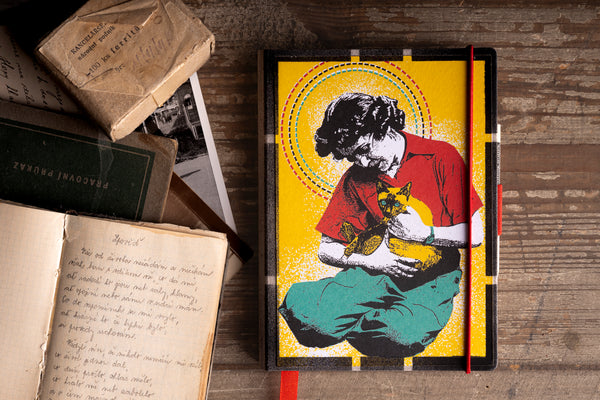 A hardcover diary with a vibrant graphic illustration of a woman holding a cat, set against a yellow background with a red elastic closure, displayed on a rustic wooden table next to vintage books.