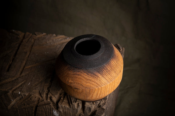 A small round wooden vessel or vase with a dark charred top and a warm honey-brown body, displayed on a rustic wooden block against a dark textured background.