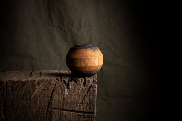 A handmade wooden ball-shaped vessel with a dark painted top and base, displayed on a rustic wooden block against a dark textured background. Made from red oak. 