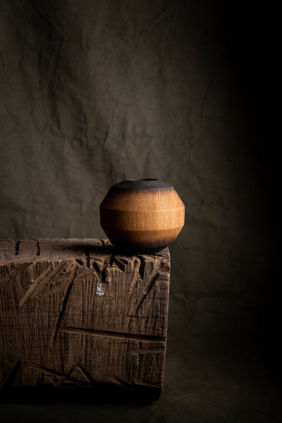 A small round wooden vase from red oak with a dark top and base sits on a rough wooden block against a dark textured backdrop.