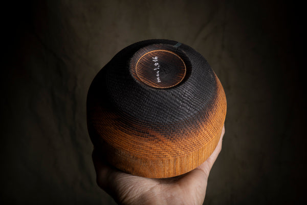 A hand holds a round wooden vessel photographed against a dark, textured background.