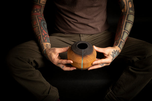 A person with heavily tattooed arms holds a small handcrafted wooden hollow form with a dark top and a thin light blue stripe, set against a solid black background. Hand turned in Prague from red oak. 