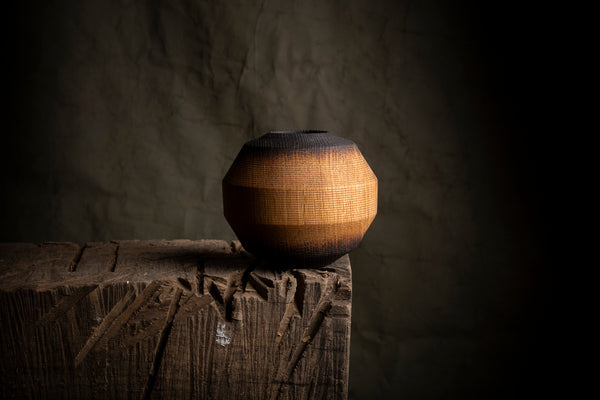 A handmade wooden vase with a textured, hand-hammered finish and a dark charred top, displayed on a rustic wooden block against a dark textured background.
