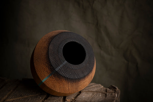 A handmade wooden hollow vessel with a dark  top and rim, resting on a rustic wooden surface against a dark, textured backdrop.