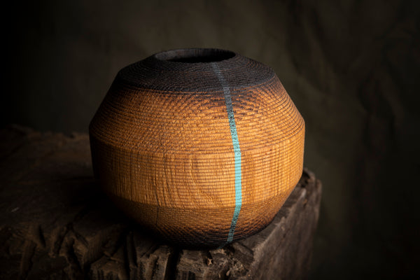 A round wooden hollow vessel with a rounded, bulbous shape, featuring a textured pattern of orange, brown, and black with a vertical light blue stripe, displayed on a rustic wooden stump against a dark background.