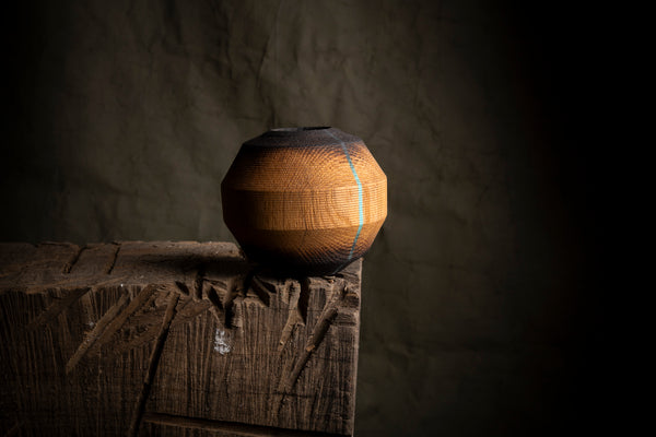 A faceted wooden decorative vase with a warm honey-brown finish made by tung oil and a thin light blue stripe, displayed on a rustic wooden block against a dark textured background.