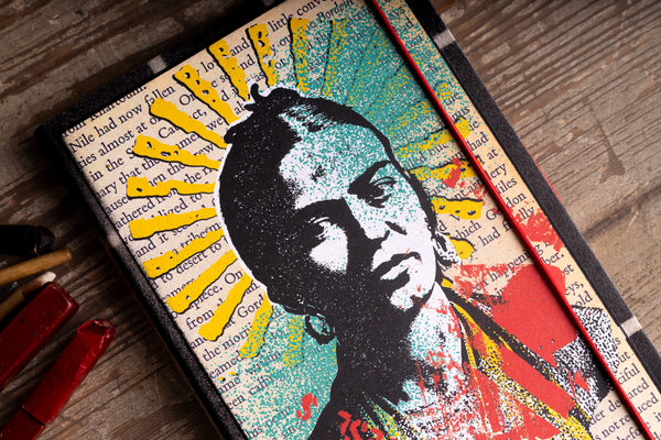 A hardcover notebook with a stylized portrait of a woman's face with a black silhouette overlay, set against a background of teal, red, yellow, and cream book text. The canvas is shown on a rustic wooden table next to colored pencils.