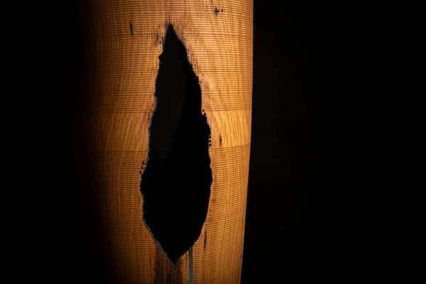 A close-up of a cylindrical vase with a prominent irregular  hole, showing a warm golden-brown textured surface of oiled red oak against a solid black background.