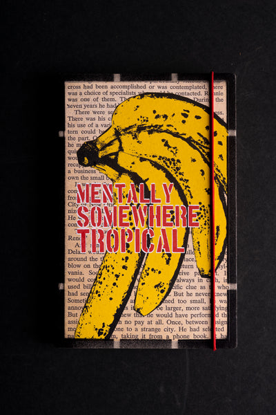 A notebook cover titled 'MENTALLY SOMEWHERE TROPICAL' featuring a large yellow banana illustration over a book page background, photographed on a dark textured surface.