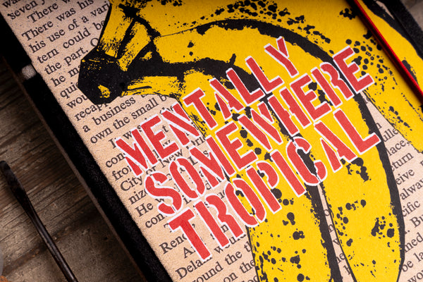 A close-up of a yellow graphic print on a notebook featuring a yellow banana and bold red text reading 'MENTALLY SOMEBWHERE TROPICAL', placed over a book page on a wooden surface.