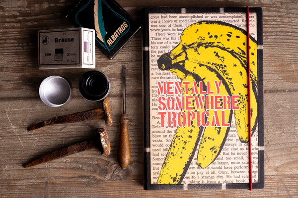 A notebook with a black cover, a yellow banana illustration, and the text 'MENTALLY SOMETHERE TROPICAL' in red and white, styled on a rustic wooden table with vintage tools.
