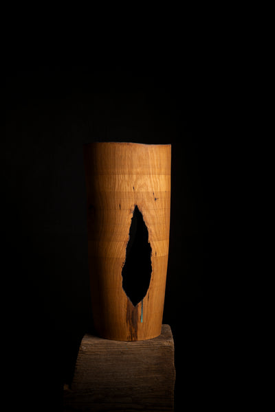 A rustic wooden cylinder vase with a prominent natural crack, displayed on a dark wooden block against a solid black background.