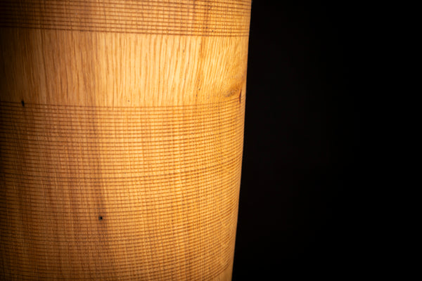 A close-up of a cylindrical wooden object with a warm golden-brown finish and visible wood grain, set against a solid black background. Also visible on this photo is textrure made on the lathe on the vessel turned from red oak. 