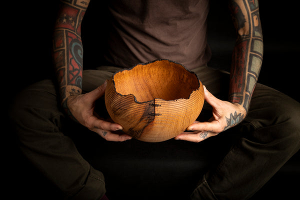 A person with tattooed arms holds a handcrafted wooden bowl from red oak with a raw, rustic edge, shown against a dark background.