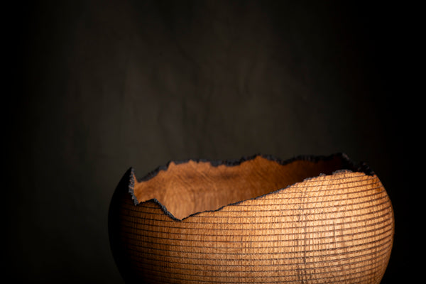 A close-up of a textured wooden vase with a jagged, charred edge, illuminated from within against a dark, moody background. The vase is made from red oak. 