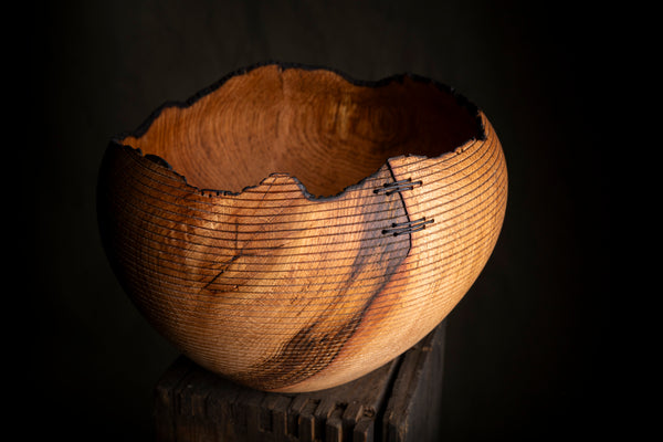A rustic handcrafted wooden bowl with a raw, burnt-edge rim and fine horizontal line detailing on the exterior, displayed on a dark wooden block against a black background. The bowl was turned on the lathe from red oak. 