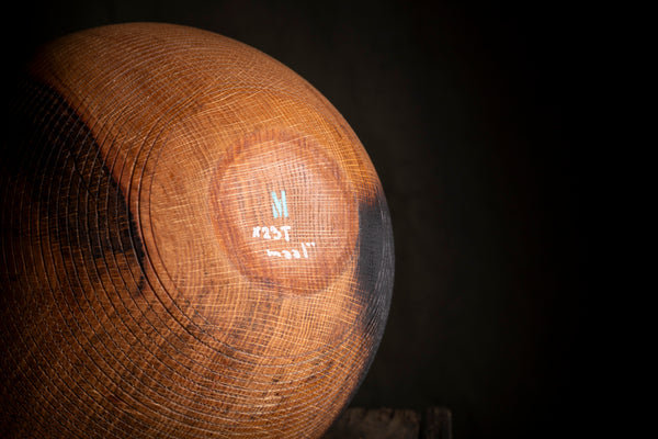 A close-up of a bottom of a round wooden object, showing a prominent natural wood grain pattern and a small white logo in the center, set against a dark background.