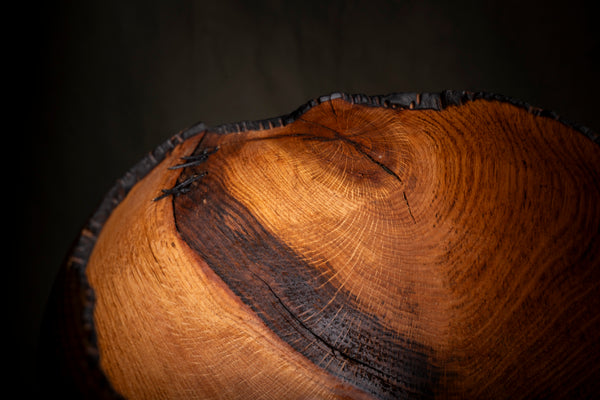 Close-up of a large wooden bowl  with a natural dark charred edge, showing rich warm brown wood grain and dark charred streaks on a dark background.