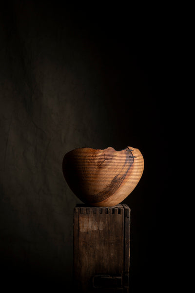 A carved wooden bowl with a distinctive broken-edge design, displayed on a rustic wooden pedestal against a dark, moody background.