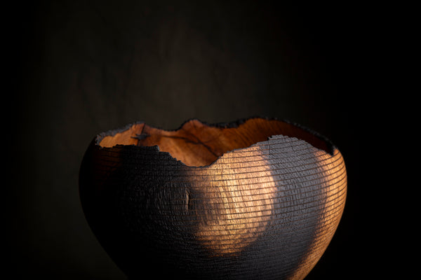 A dark, textured wooden bowl with a rough, basket-like exterior and a smooth, warm brown interior, photographed against a very dark background with dramatic light highlighting its texture.