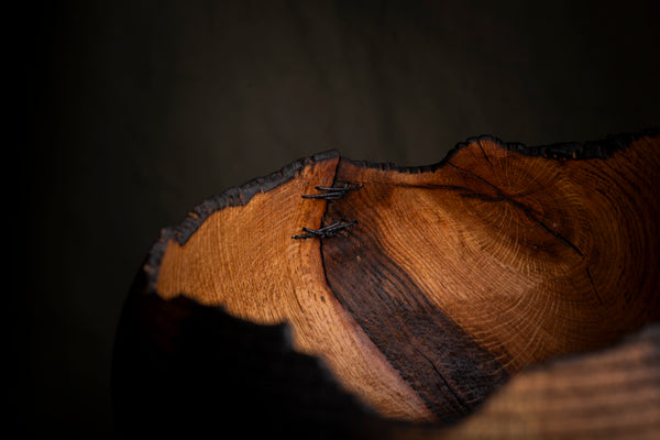 A close-up of a rustic wooden bowl  with a thick natural charred edge, showing rich warm brown wood grain and dark charred edges against a dark background. Also visible are the stitches securing the crack by the opening. 
