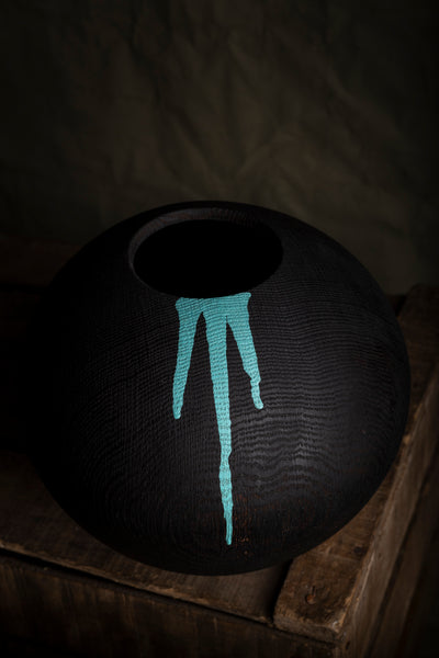 A black textured wooden vessel made from red oak with a turquoise painted dripping design, resting on a rustic wooden surface against a dark background.