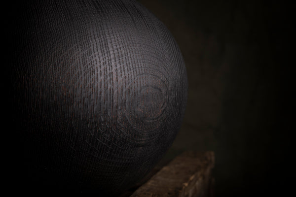 A close-up of a dark, textured spherical object with a visible wood grain, resting on a wooden surface against a dark background.