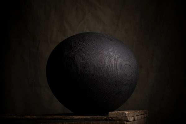 A large spherical black  wooden decorative object with a textured  wood grain surface sits on a rustic wooden pedestal against a dark, moody background.