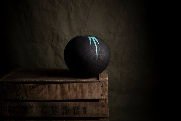 A matte black spherical decorative object  made from red oak with a turquoise painted vertical splashes, displayed on top of a rustic wooden crate against a dark textured background.