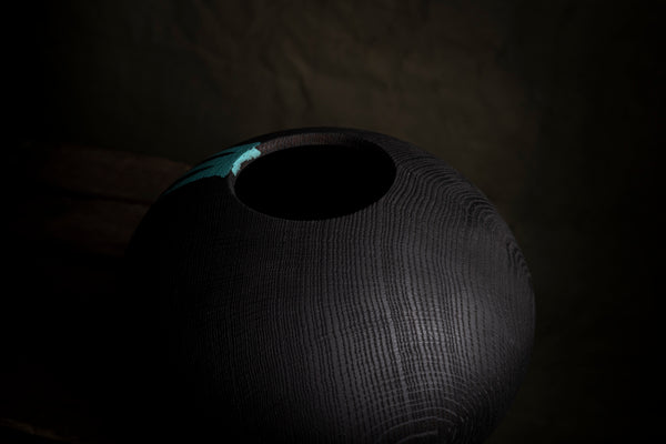 A dark textured wooden vessel  with a circular opening, shown in dramatic low light against a dark background. The object is made from red oak.