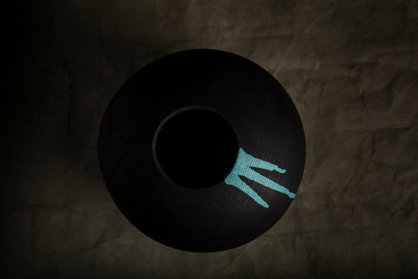A black textured round hollow form from red oak with a hole in the center, photographed from above on a dark brown crumpled-paper background. A small turquoise woven accent is visible on the right side of the rim.