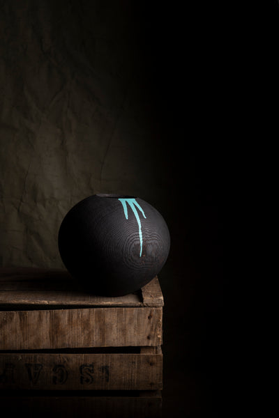 A matte black spherical decorative object with a light blue paint splashes, displayed on stacked wooden crates against a dark textured background.