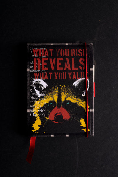 Handmade notebook "Little Racoon" with a screen-printed cover of a raccoon face in yellow, red and black under the red stencil words "WHAT YOU RISK REVEALS WHAT YOU VALUE", with white typewriter text on the left. It has a black textured border, a red elastic closure, a red ribbon bookmark and a pen loop, on a black background.