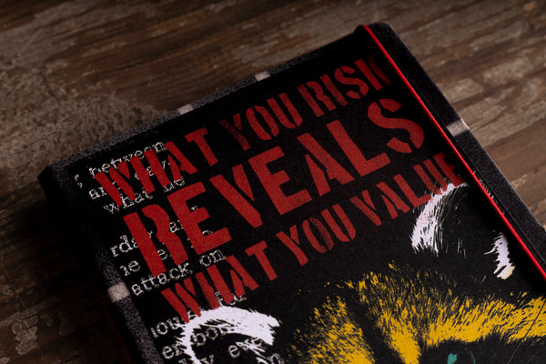 Close-up of the top of the "Little Racoon" handmade notebook cover, showing the red stencil lettering "WHAT YOU RISK REVEALS WHAT YOU VALUE" over white typewriter text, the raccoon's ears and the textured black border, on a wooden surface.