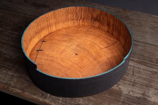 A round wooden bowl with a dark textured exterior and a bright teal painted rim, shown on a rustic wooden tabletop. The bowl is made from red oak in the Prague based studio called Maalstrom studio. 
