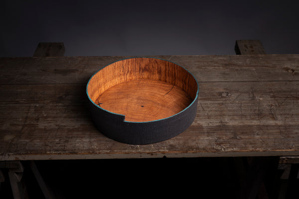 A round wooden bowl with a painted blue rim, displayed on a rustic wooden workbench against a dark gray background.