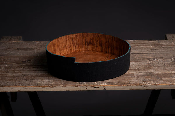A circular wooden bowl from red oak with a black rim sits on a rustic wooden workbench against a dark black background.