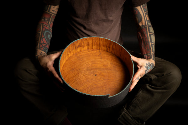 A person with tattooed arms holds a round wooden hand turned bowl from red oak against a dark background. The product is shown in close-up, with the warm orange-brown wood grain interior and a dark exterior rim visible.