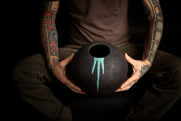 A person with heavily tattooed arms holds a matte black spherical vessel  from red oak with a turquoise drip design against a dark background.