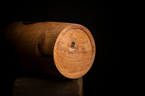 A close-up of a bottom of a tall wooden vessel, showing a warm brown wood grain surface of red oak with a small black and white logo, set against a solid black background.