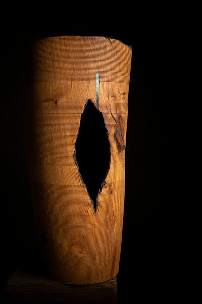 A cylindrical wooden object with a warm honey-brown finish and a prominent dark, irregular hole  on the front, photographed against a solid black background.