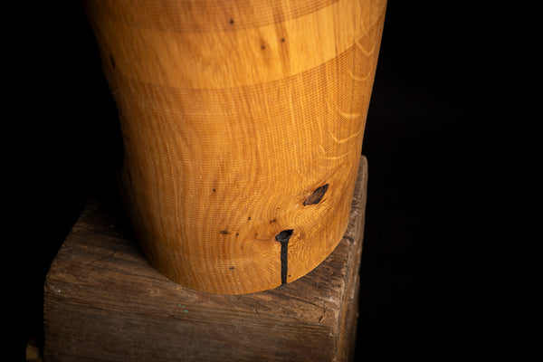 A close-up of a cylindrical wooden object with visible wood grain and a small dark hole near the bottom, resting on a rough wooden block against a black background.