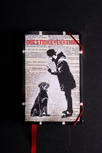 A hardcover handmade notebook or journal with a black and white graphic print of a person looking at a dog, with the red text 'QUESTION EVERYTHING' across the top, photographed against a dark background with a red ribbon bookmark.