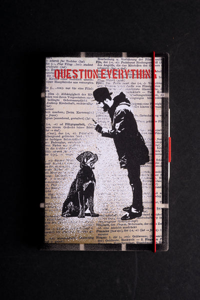 A hardcover notebook  with a black and white graphic illustration of a man and a dog over a vintage dictionary page background, featuring the red text 'QUESTION EVERYTHING' on the cover, a red elastic closure, and a black spine, shown on a dark background.