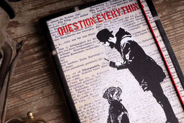 A handmade notebook featuring a black-and-white stencil-style illustration of a person and dog over a dictionary page background, with the word 'QUESTION EVERYTHING' in red across the top, displayed on a rustic wooden surface.