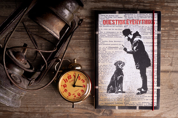 A hardcover journal titled 'QUESTION EVERYTHING' lies on a rustic wooden table alongside a vintage brass alarm clock, a weathered lantern, and a black-and-white silhouette illustration of a person and  a dog.